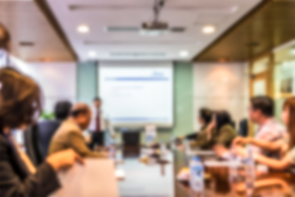 out of focus image showing a group of people around an elongated boardroom table looking at a presentation being shown on the far wall.