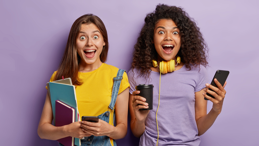 Two young students standing side by side holding text books and a cell phone showing joy and excitement.