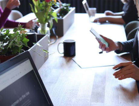 Three people are seated around a counter with a row of plants down the centre of the counter. the people are all working individually either on laptops or completing documents while referring to their cellphone.