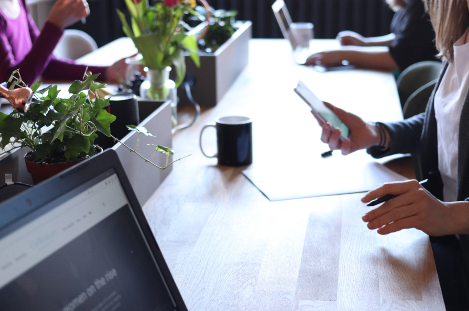 Three people are seated around a counter with a row of plants down the centre of the counter. the people are all working individually either on laptops or completing documents while referring to their cellphone.