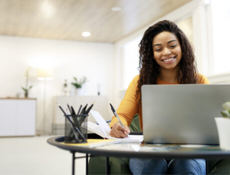 Woman sitting at desk, using computer and writing in notebook
