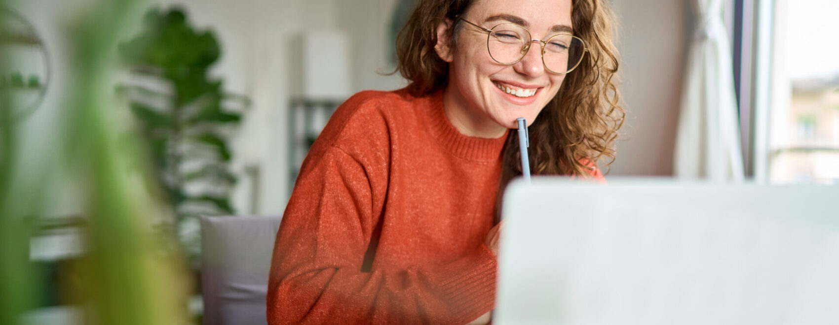 A photo of a person in an orange sweater with glasses sitting at a desk, looking at her laptop.