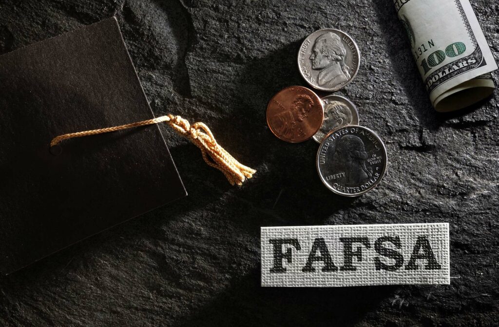Stock photo of black graduation cap with coins, a $100 bill and a slip of paper with the word FAFSA on it