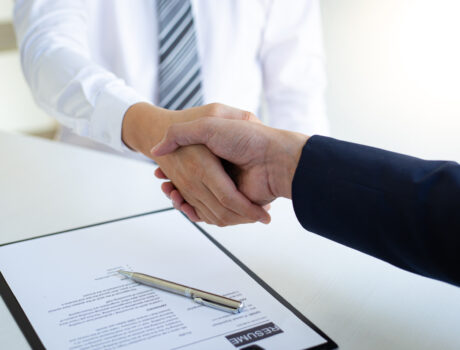 photo of two peoples arms performing a handshake; clipboard with a resume on desk